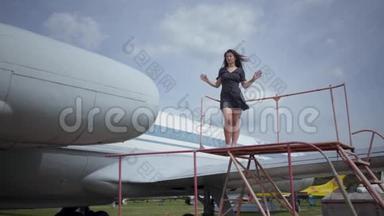 快乐的年轻微笑的黑发女孩看着相机站在白色的大飞机触摸她的头发。 夏季